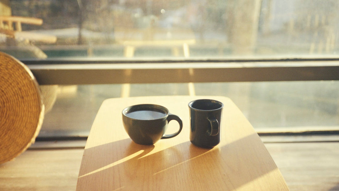 Two dark coffee cups on a sunlit wooden table beside a window, reflecting a quiet moment shared.