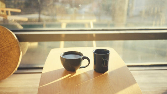 Two dark coffee cups on a sunlit wooden table beside a window, reflecting a quiet moment shared.